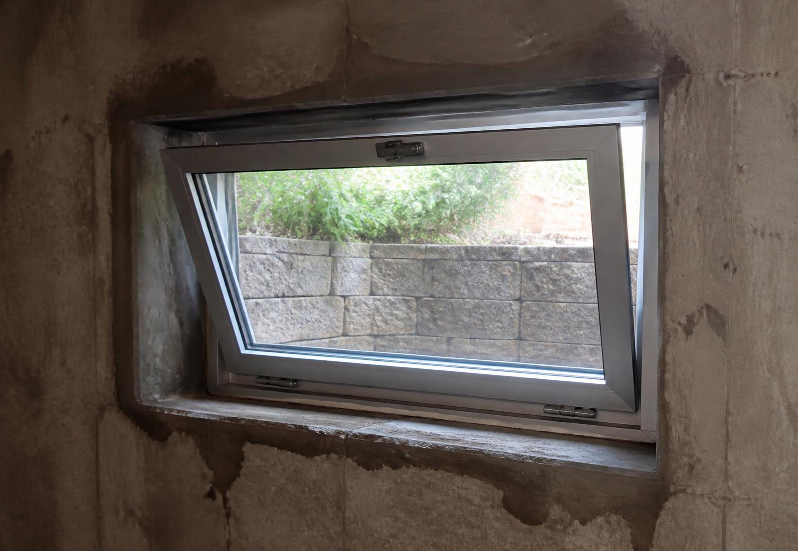 Aluminum hopper window tilted inward from its sill hinge in a concrete basement wall